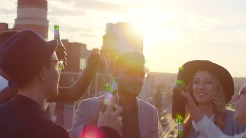 Group of mixed raced young men and women smiling, dancing and toasting with beer bottles while having outdoor party on rooftop at sunset
