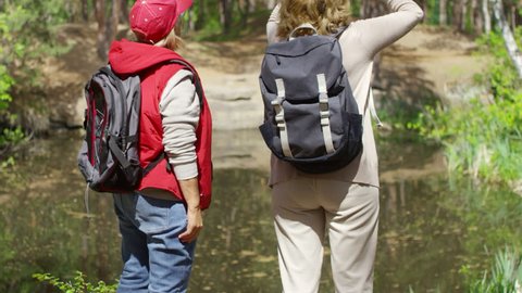 Tilt up shot of curious elderly women with backpacks standing by the pond in forest and looking up through binoculars
