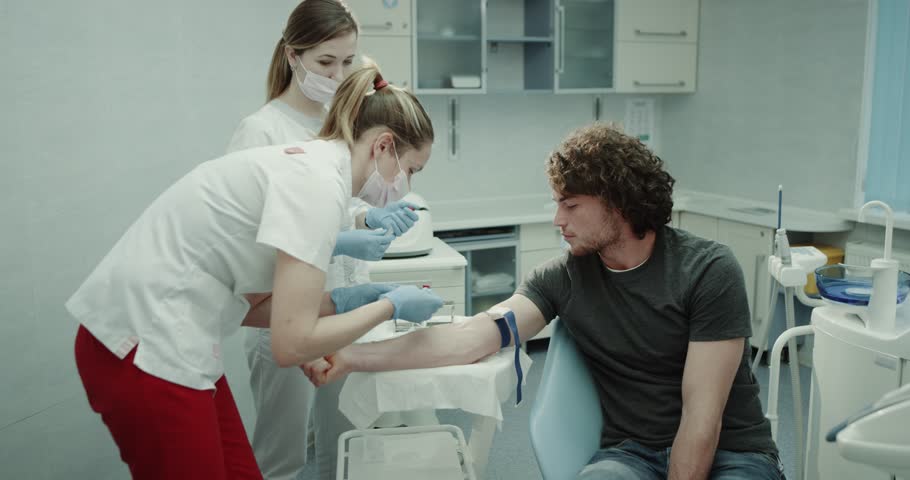 Doctor and her assistance make a blood test of a young donor , collecting the blood from hand , man sitting on the medical chair in a modern clinic. | Shutterstock HD Video #1021635298