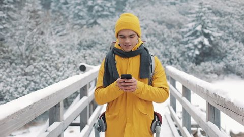 A young tourist standing on the bridge and using smartphone.Winter time. Beautiful mountains and snow-covered lake background