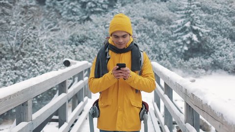 A young tourist standing on the bridge and using smartphone. Hiker reading good news celebrating looking at cell phone. Winter time. Beautiful mountains background