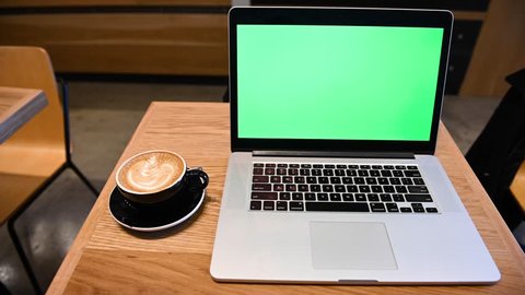 Laptop with green screen on a coffee shop table with a black cappuccino coffee cup 