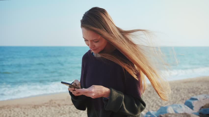 Female with a smartphone or mobile phone is texting or writing a letter to beloved friend or couple using 4g social network app or application from the beach in harmony moment wile wind is waves hair Royalty-Free Stock Footage #1021647568