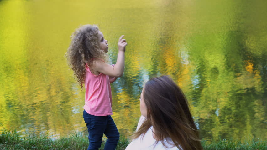 Family picnic by lake. Mother and daughter outdoors. Curly cute little girl holding red apple and telling story. Woman is surprised.