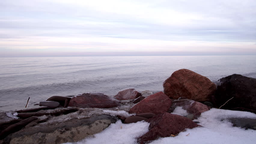 Snowy shore with big, iced-covered orange rocks, stones by calm, tranquil Lake Superior in winter, Upper Peninsula, Michigan. Looping 4K UHD clip, suitable for motion backgrounds