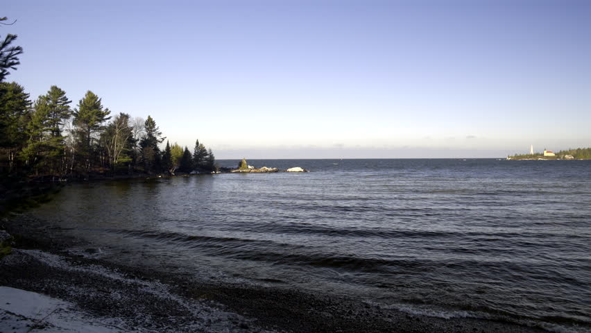 Lake Superior winter shoreline with conifers, gentle fresh-water waves and lighthouse in background, Upper Peninsula, Michigan. 4K UHD