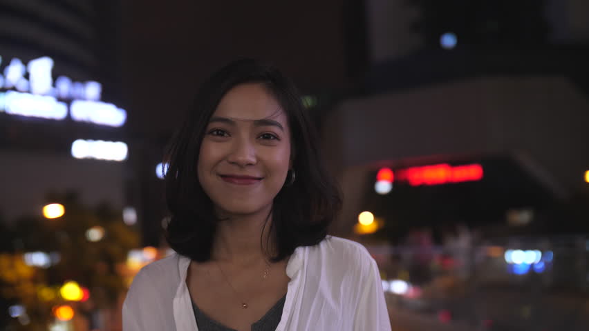 Slow motion of one beautiful young Chinese girl looking at camera smile in the summer night with wind blowing her hair city traffic and urban neon light at background,attractive young woman footage 4k