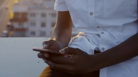 Tilt up shot of young african american man sitting on urban rooftop in the evening, watching video on smartphone and smiling