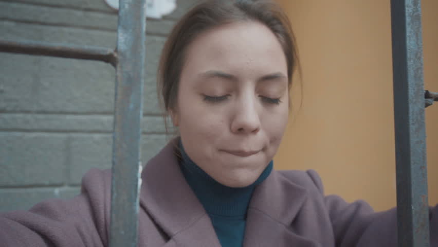 The girl in the lilac coat stands behind the fence on the street and looks