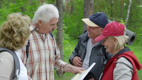 Medium shot of happy elderly women and men with backpacks laughing and talking while looking at paper map and deciding which forest trail to hike