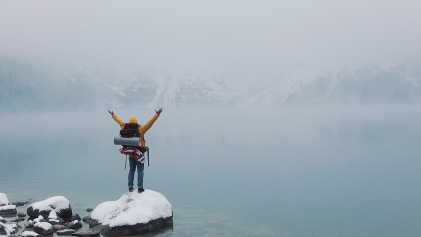 American hiker stands on a stone near a mountain lake and raising hands on mountain top winner concept. Snowy blurred mountains cape and lake