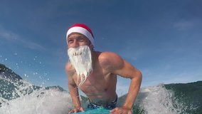 SLOW MOTION CLOSE UP: Cheerful Santa surfer paddling and catching fun waves in sunny ocean. Smiling Santa Clause surfer riding waves towards the shore to wish a Merry Christmas. Santa Clause surfing - Powered by Shutterstock - Get 15% off with code: PIKWIZARD15