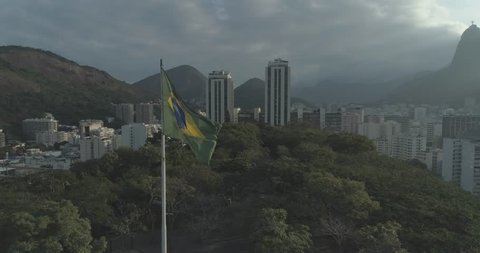 Aerial drone shot circling waving sunlit Brazil national flag in forest with Christ the Redeemer and city buildings in background, Rio de Janeiro