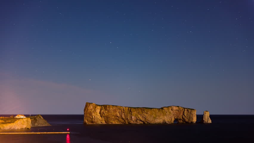 Timelapse, time lapse of Rocher Perce rock in Gaspe Peninsula, Quebec, Gaspesie region, Canada at dark night with starry sky, stars at Saint Lawrence gulf, wharf, dock, pier, lighthouse, reflection