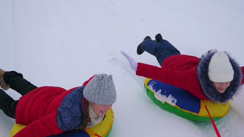 Girls in red jackets ride through snow in winter on an inflatable snow pipe and play super heroes. happy girls relaxing in winter park for christmas holidays. Slow motion | Shutterstock HD Video #1021732174