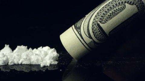 View of white powder drug being snorted up rolled up dollar bill on black reflective table top.