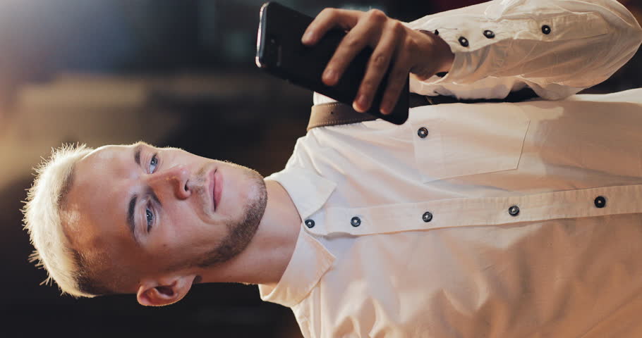 Vertical video. Happy young attractive man using smartphone standing on street of night town. Vertical frame for mobile devices