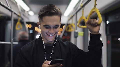 Portrait of attractive man in headphones holds the handrail, listening to music and browsing on mobile phone in public transport. City lights background