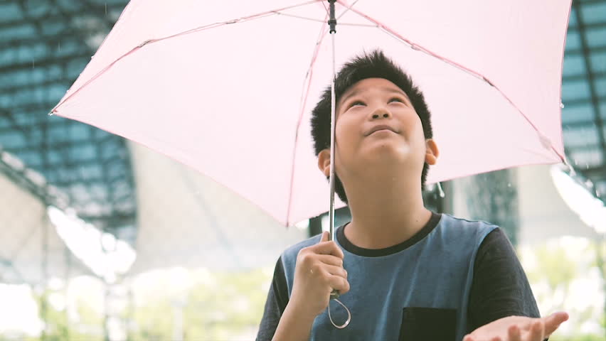 Slow motion of Asian boy with toothy smile holding pink umbrella and catching raindrops during heavy rain