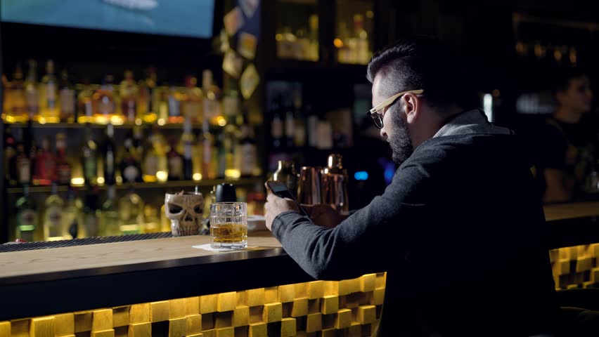 Behing shot of a man quietly sitting at the bar with glass of whiskey, chatting online.