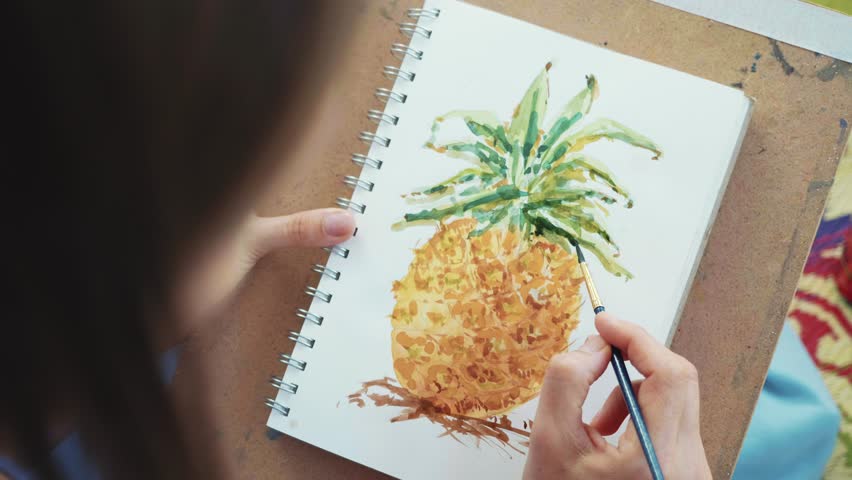 Close-up of female hand with paintbrush is drawing pineapple in sketchbook by watercolors