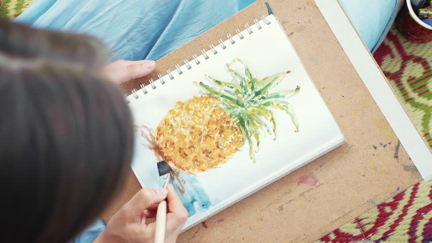 Close-up of female hand with paintbrush is drawing pineapple in sketchbook by watercolors