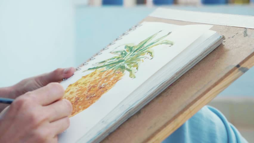 Close-up of female hand with paintbrush is drawing pineapple in sketchbook by watercolors