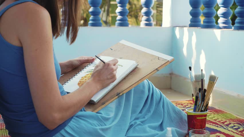 Young female artist painting a pineapple by watercolors. Woman sitting on the floor and drawing in sketchbook. 