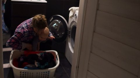 Woman takes out the laundry from the washing machine. She holds her baby in her arms. It is difficult for her to cope alone. mother's day, single mother