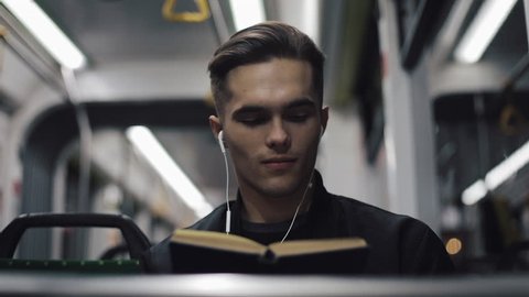 Young handsome man sitting on public transport reading a book - commuter, student, knowledge concept. Young man with headphones in the tram reading a book