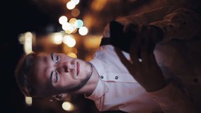 Vertical video. Men send voice message using his smartphone on the street of the night city. Businessman using mobile phone for business communication voice message