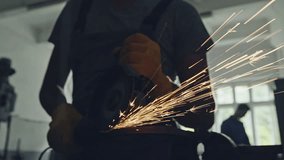 A close up of man using grinder in dark workshop and throwing bright orange sparks - slow motion