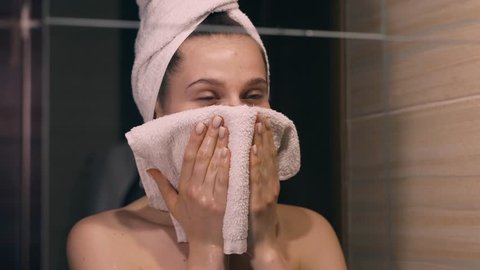 Close-up girl wipes her face with a towel. Facial treatment. Beautiful woman after a shower. The girl stands in the bathroom in a towel. Woman dries her hair with a towel and looks in the mirror