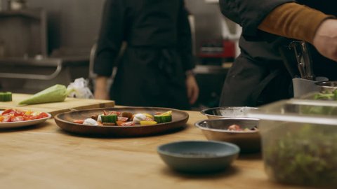 Professional chef arranging lettuce leaves on a wooden plate with other ingredients in industrial kitchen with soft interior lighting. Close up shot on 4k RED camera.