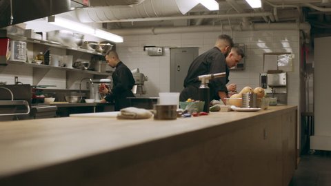 Pan of professional chefs working at their stations at a long counter in industrial kitchen with soft lighting. Medium to close up shot on 4k RED camera on a gimbal.