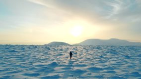 Man hiking in winter aerial. Drone video of a young man hiking snowy mountain landscape at beautiful winter sunset. Male mountaineer with trekking poles and a backpack. Travelling in wintertime