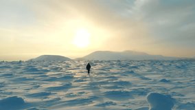 Man hiking in winter aerial. Drone video of a young man hiking snowy mountain landscape at beautiful winter sunset. Male mountaineer with trekking poles and a backpack walking on mountain ridge .