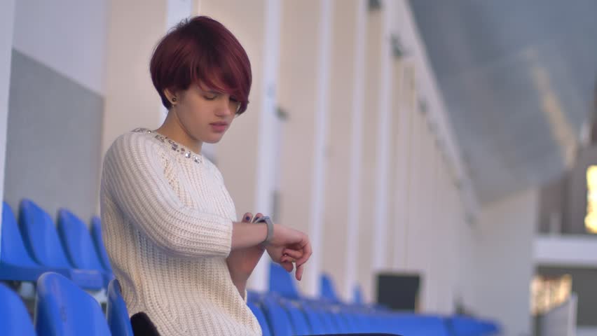 Portrait of happy beautiful pink-haired girl sitting on stadium and looking at watch.