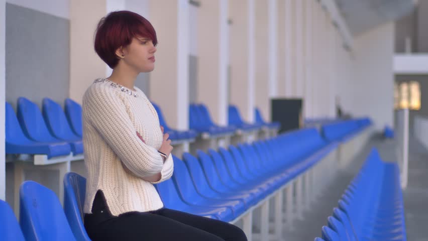 Portrait of bored beautiful pink-haired girl sitting on stadium and waiting for something with arms crossed.