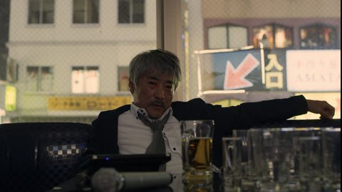 Drunk sad Japanese man sitting at a booth in a karaoke room alone with soft interior lighting. Medium shot on 4k RED camera.