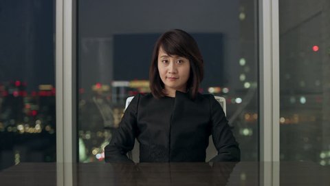 Professional Japanese woman sitting at a conference table in front of a large window with a view of night time in a conference room with soft interior lighting.