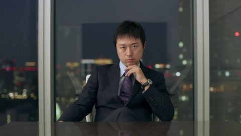 Contemplative Japanese man sitting at a conference table in front of a large window with a view of night time in a conference room with soft interior lighting.