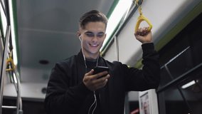 Portrait of attractive man in headphones holds the handrail, watching video on smartphone in public transport. City lights background