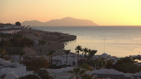 Sunrise over the red sea illuminates the coast of Sharm El Sheikh. Tourist resort with palm trees and buildings near the coastline
