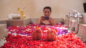 Young Woman Having Fun in Bath with Floating Red Flowers and Taking Video with Smartphone. Girl Playing With Her Feet in Spa. 4K Slowmotion. Bali, Indonesia.