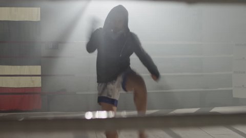 Muay Thai fighter in hoodie throwing punches at camera, shadow boxing, training in the middle of a foggy boxing ring backlit with flares in the background