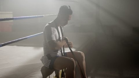 Muay Thai fighter puts on hand wraps while sitting on the edge of a boxing ring in a musty boxing gym, camera rotates around from side to front స్టాక్ వీడియో