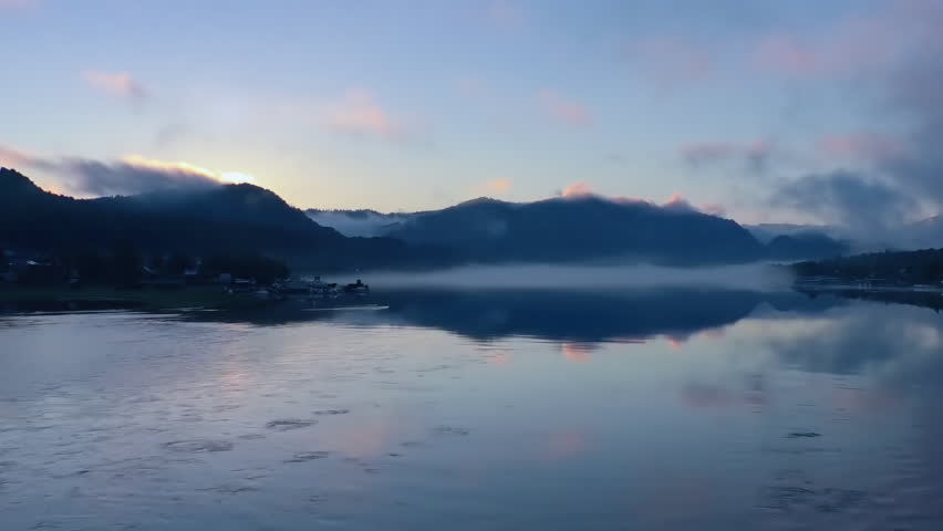 Sunrise over the mountain lake. Early summer morning. Bright clouds fly over the tops of the mountains, overgrown with forest. Swirling fog. Flowing water. The water reflects clouds.