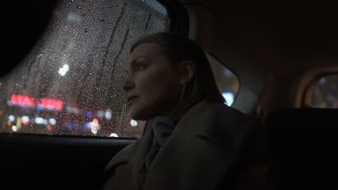 Sad woman riding taxi in evening, looking into car window on rain, city lights