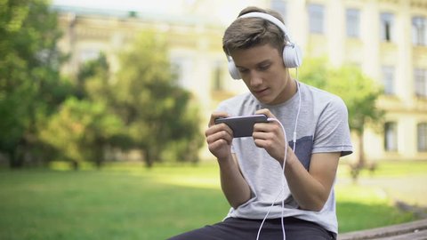 Gamer in headphones sitting in park, playing game on mobile phone, addiction